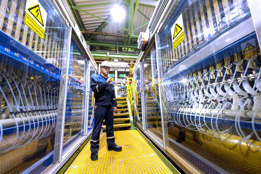 Ein Fabrikmitarbeiter in Arbeitskleidung steht in einem Raum voller Glasschränke, hinter denen sich eine Vielzahl an Kabeln und Schläuchen befinden.