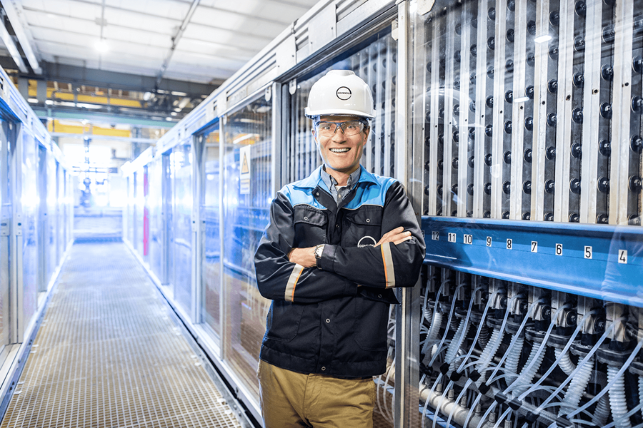 Ein Mitarbeiter in Arbeitskleidung steht freundlich lächelnd mit verschränkten Armen in einem Raum voller Glasschränke, hinter denen sich eine Vielzahl von Kabeln und Schläuchen befindet.