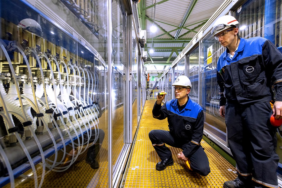 Zwei Mitarbeiter mit Schutzbrillen und Arbeitskleidung stehen in einem Raum innerhalb eines Kraftwerks und begutachten Kabel und Schläuche, die hinter Glas geschützt sind.