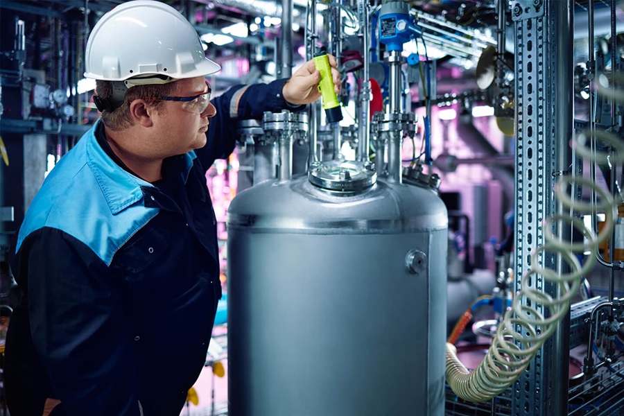 Ein Fabrik-Mitarbeiter in Arbeitskleidung und mit Schutzhelm und -Brille hält eine gelbe Taschenlampe in der Hand und inspiziert eine Maschine.