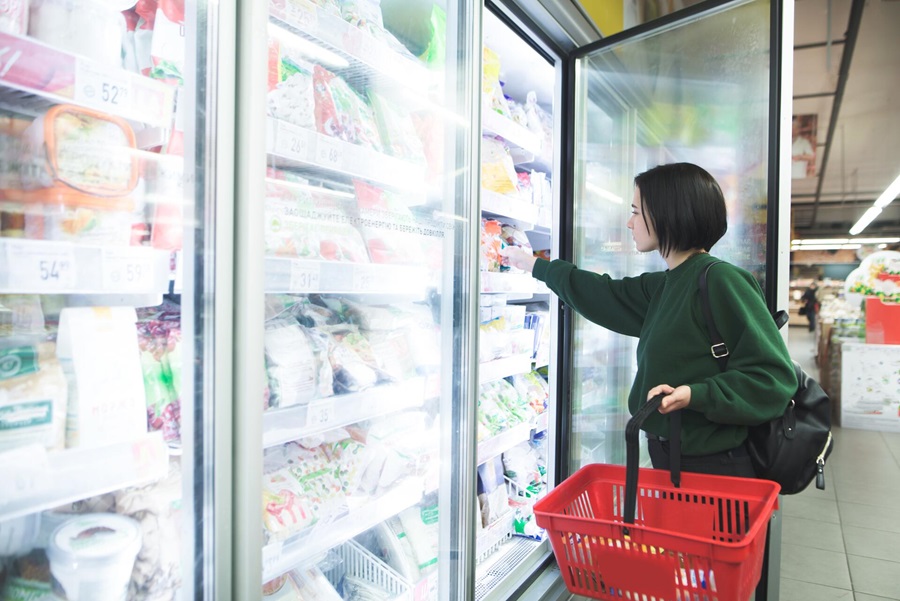 Eine Frau steht in einem Supermarkt und greift nach den Lebensmitteln in einem geöffneten Kühlschrank.