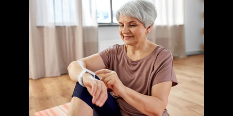 Eine ältere Frau sitzt in ihrer Wohnung und bedient ihre Smart-Watch.