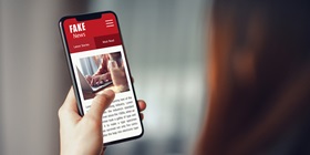 A smartphone displays a news site with a large headline about fake news, along with typical website elements like images and menus.