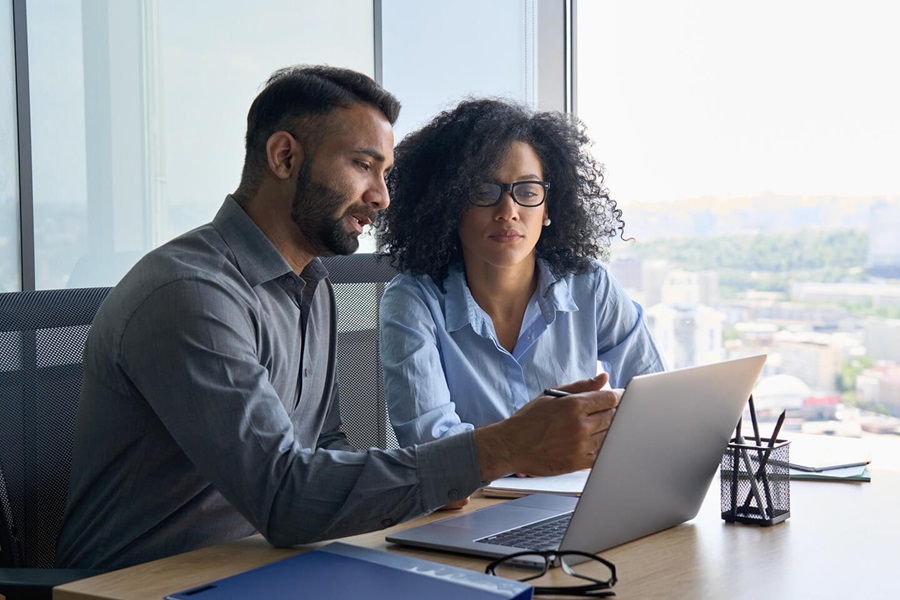 Zwei Fachleute prüfen Arbeit am Laptop am Fenster, Mann zeigt auf Bildschirm, Frau schaut zu