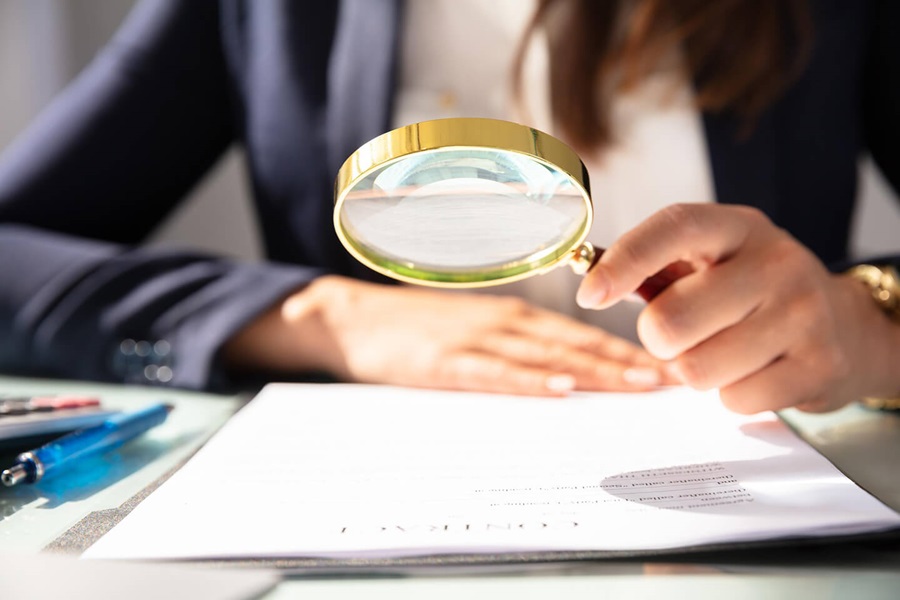 Person in Geschäftskleidung untersucht Dokument mit Lupe, Papiere auf dem Schreibtisch sichtbar